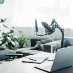 Test automation and software quality header. View of desk. On the desk is a laptop, notebook and pen, water glass. Next to the desk is a podcast mic.