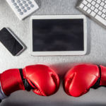 Overview of a table. A tablet, computer keyboard, and phone sit on the table. Also resting on the table is 2 red boxing gloves.