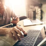 In the hero image for The Shortage of Software Engineers blog, there is a developer with multiple laptops on a table.