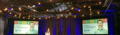 This image shows the award ceremony for the Leaders in Corporate Citizenship Award. A man stands on stage behind a podium addressing the crowd. To his left and right are two screens with Josh's photo and the KMS logo.