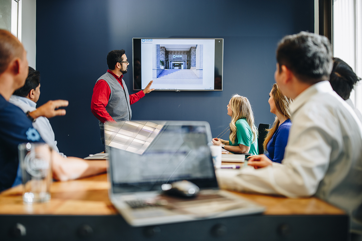 A man stands at the head of a long table, giving a presentation.