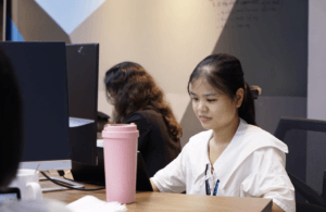 A woman sits in front of a computer working in an office. A pink coffee cup sits on her desk.